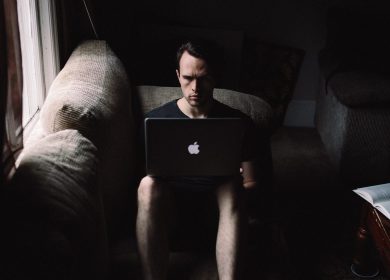Man sitting on a cough with his knees up facing the camera with a laptop on his lap