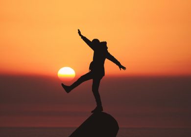 Person standing on the top of a hillpoint with arms and leg out with the sunset in the background