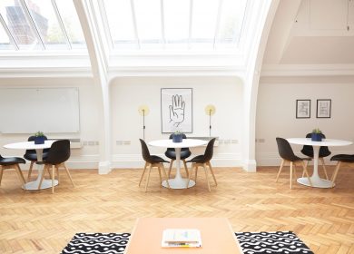Three tables with three chairs at each against a white wall, with hardwood floors and a glass ceiling