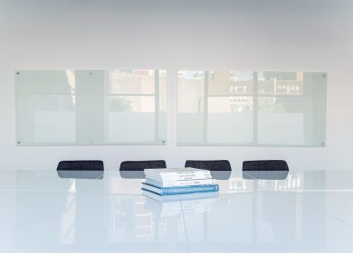 Edge of a white glass table with four chair tucked in with a glass covered wall in the background