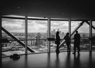 Black and white view of people in a room with large windows viewing a city in the background