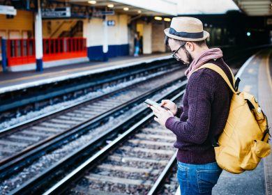 Man with hat and glasses wearing backpack standing near train tracks looking at phone