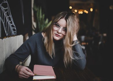 Woman with glasses and long blonde hair with hand on head and book in front of her closed with her hand in it