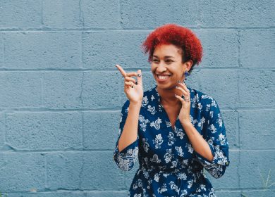 Woman standing in front of grey brick wall wearing blue shirt with read hair pointing to her right