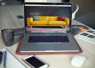 View of open laptop next to a cup of coffee, pad with pen and pencil on it, smartphone, mouse, and brochure with a chair in the background