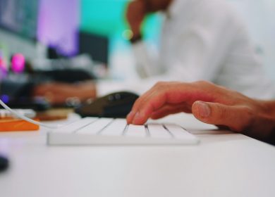 View of a keyboard and mouse with one hand on it with person in the background