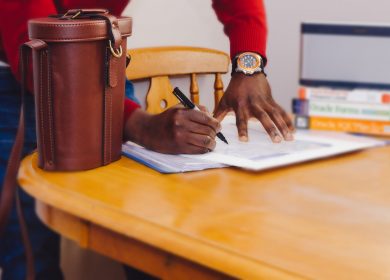 Table with Paper on it with person's hands writing on the paper and a bag on the table