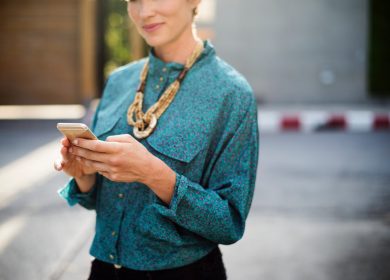 Woman standing with a smartphone in hand glancing at camera wearing teal blouse
