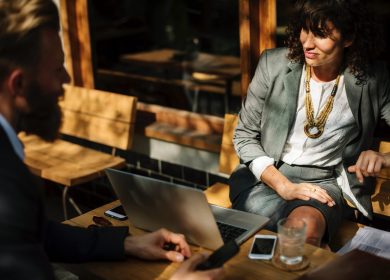 Woman facing man while both are sitting at a table with a laptop on it at a cafe