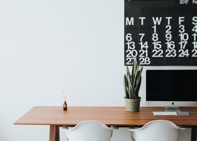 Desk with two white chairs in front of it and a computer and two plants on it in front of a calendar on the wall