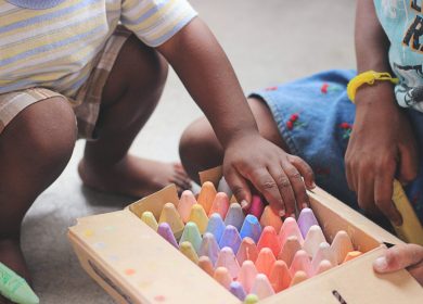 Two kids squatting down next to a container of colored chalk