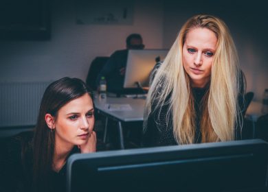 Two women looking at a computer screen with a man in the background at a desk with a computer screen also